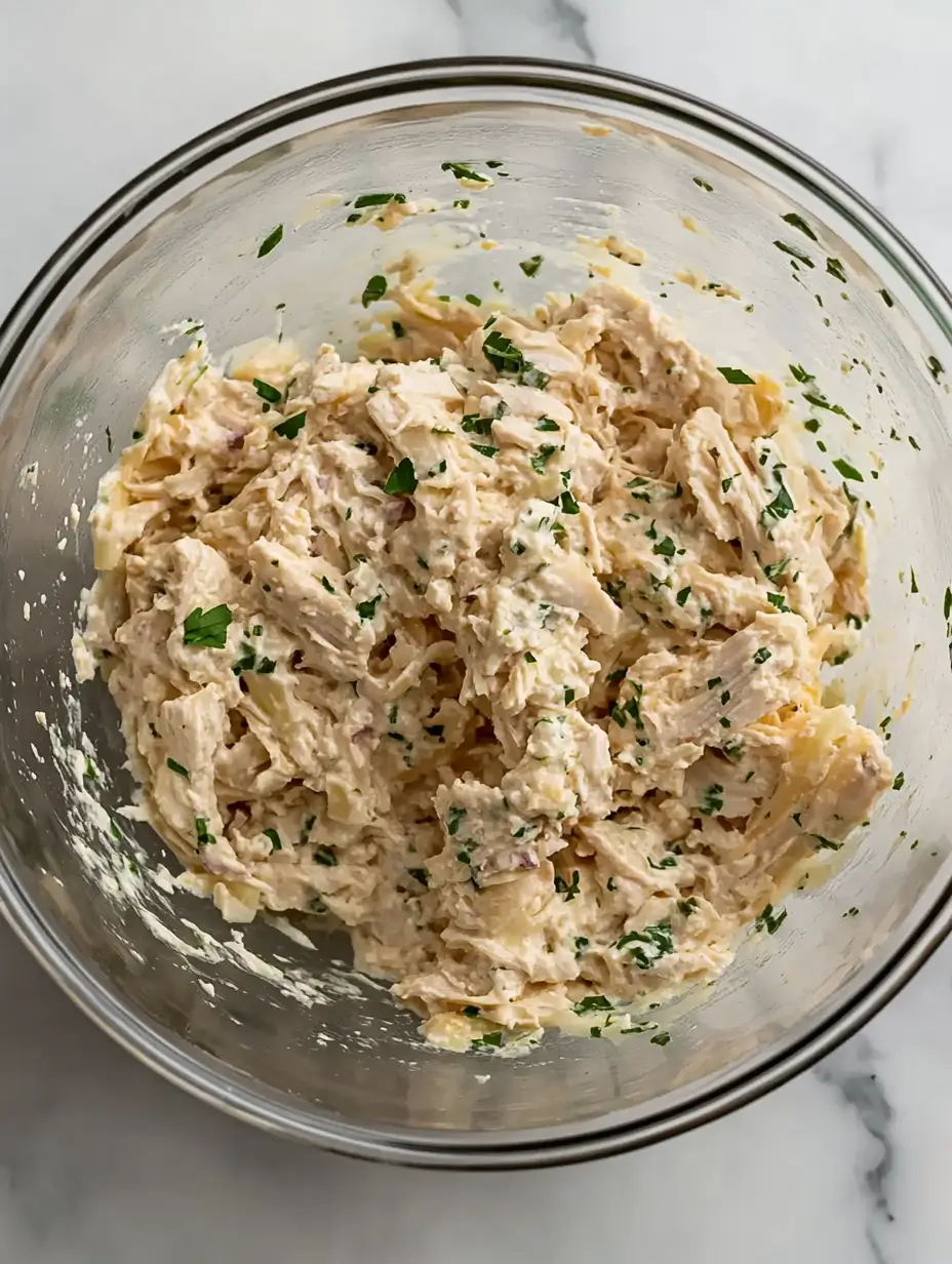 MILLION DOLLAR CHICKEN CASSEROLE 4 A glass mixing bowl containing a creamy chicken mixture with shredded chicken, cheese, and fresh parsley, fully combined and ready for the next step in the recipe.