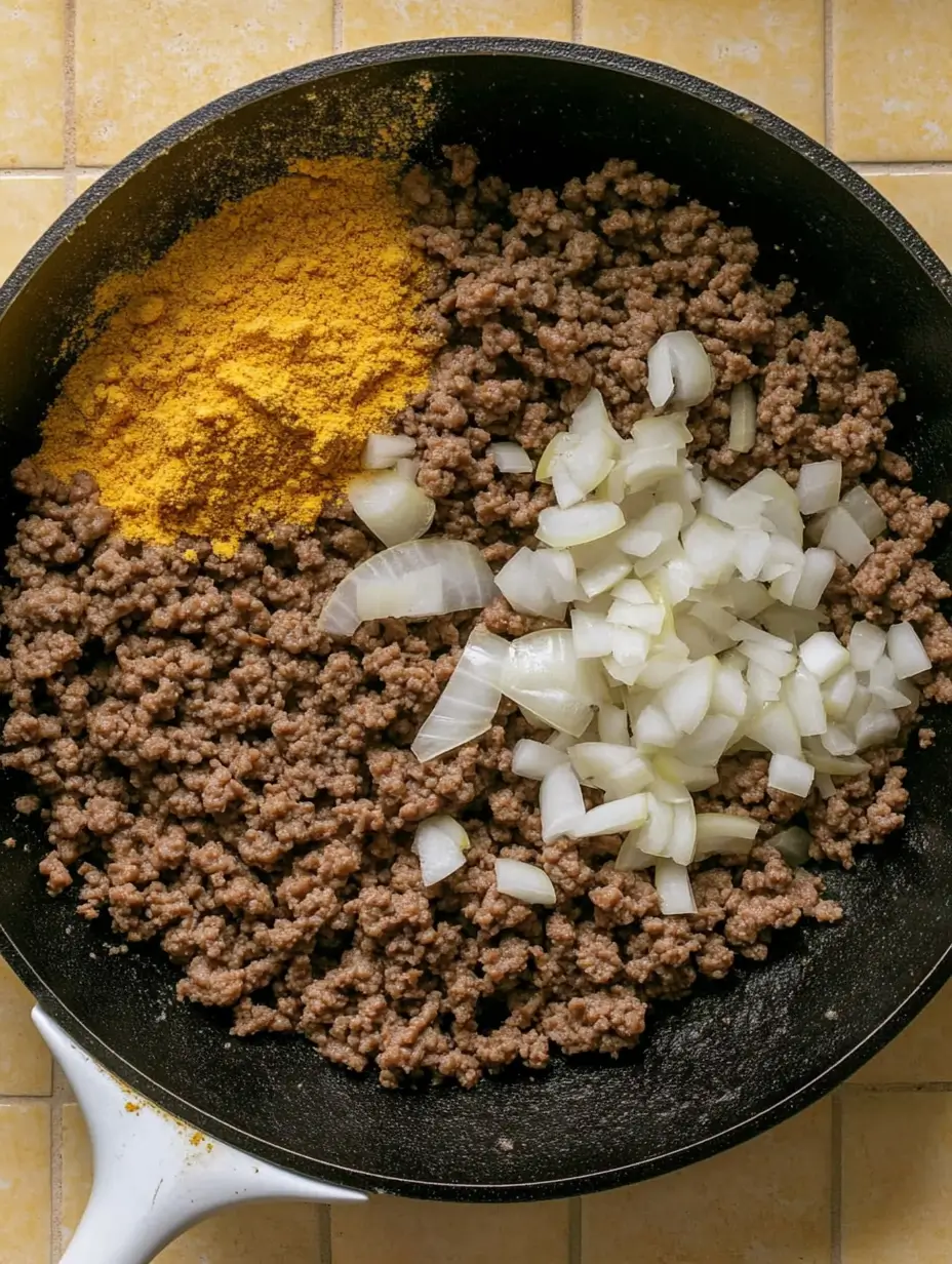 A skillet with browned ground beef, diced onions, and a pile of yellow seasoning, ready to be cooked together.