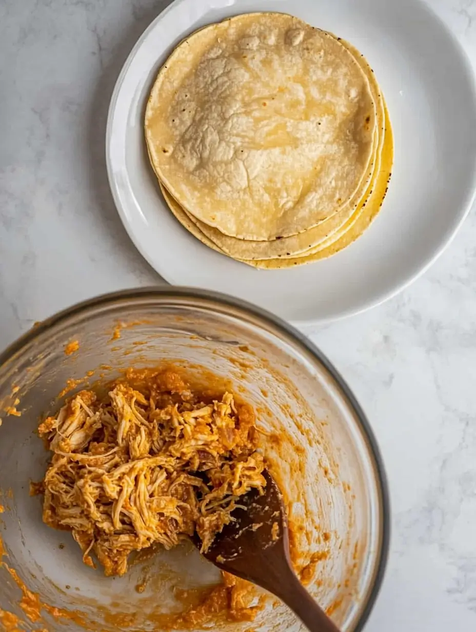 Ridiculously Good Air Fryer Flautas 16 Bowl of shredded chicken mixed with seasoning and a stack of tortillas on a white plate on a marble countertop.