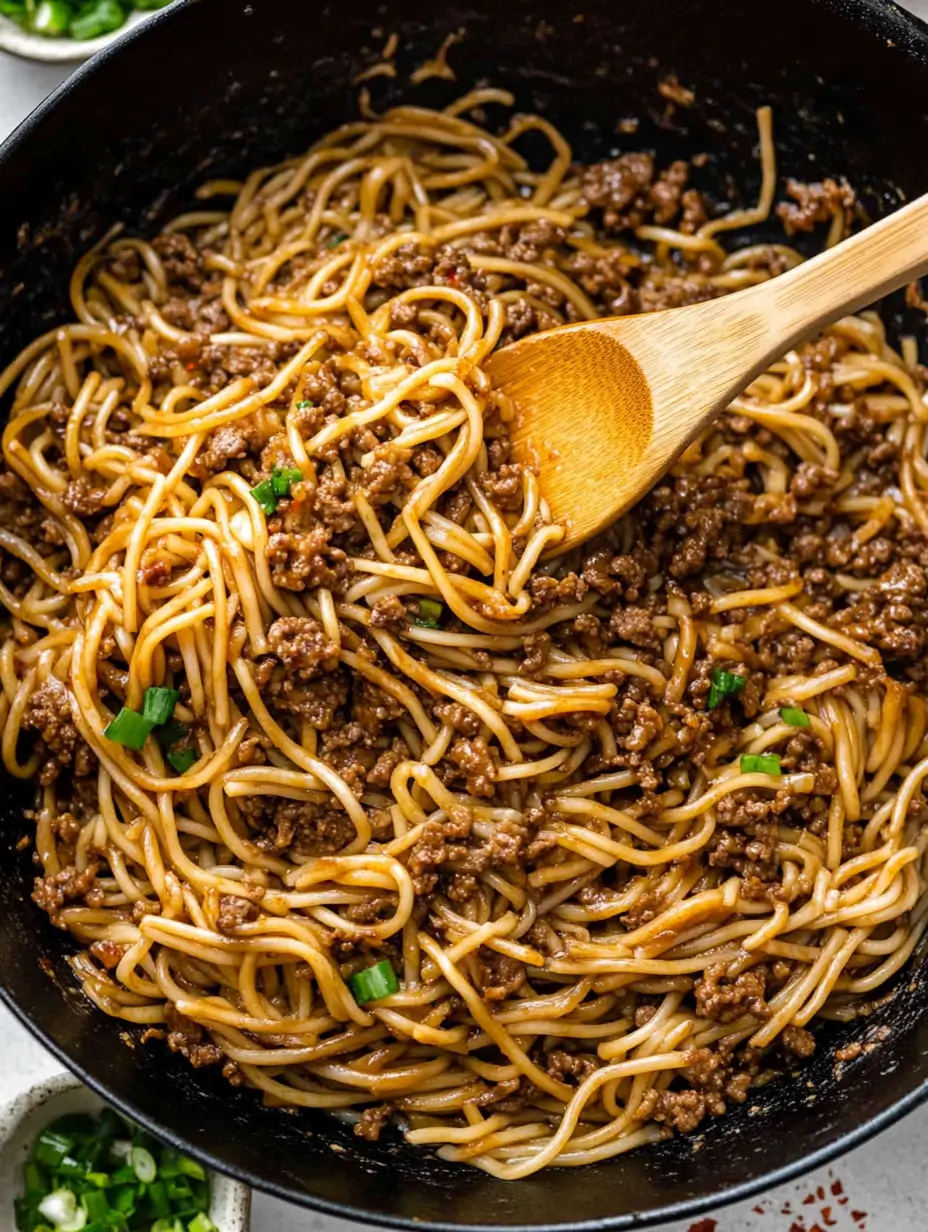 A wok filled with Mongolian ground beef noodles, stirred with a wooden spoon.