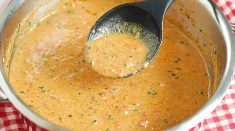 A close-up of a ladle scooping a rich and creamy soup with a reddish-orange hue, garnished with herbs, from a saucepan.