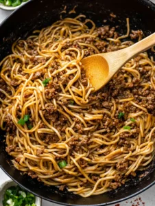 A wok filled with Mongolian ground beef noodles, stirred with a wooden spoon.
